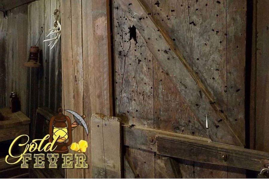 Gold Fever escape room: a weathered wooden cabin door riddled with bullet holes, with the Gold Fever logo in the corner